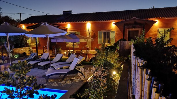A pool area with lounge chairs and umbrellas, a small house with a tiled roof, and a bamboo fence. - Villa Maria (Kefalonia)