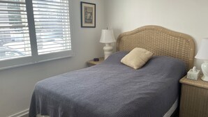 Ein Schlafzimmer mit Bett, Nachttisch, Lampe, einem gerahmten Bild an der Wand und einem Fenster mit Jalousien.