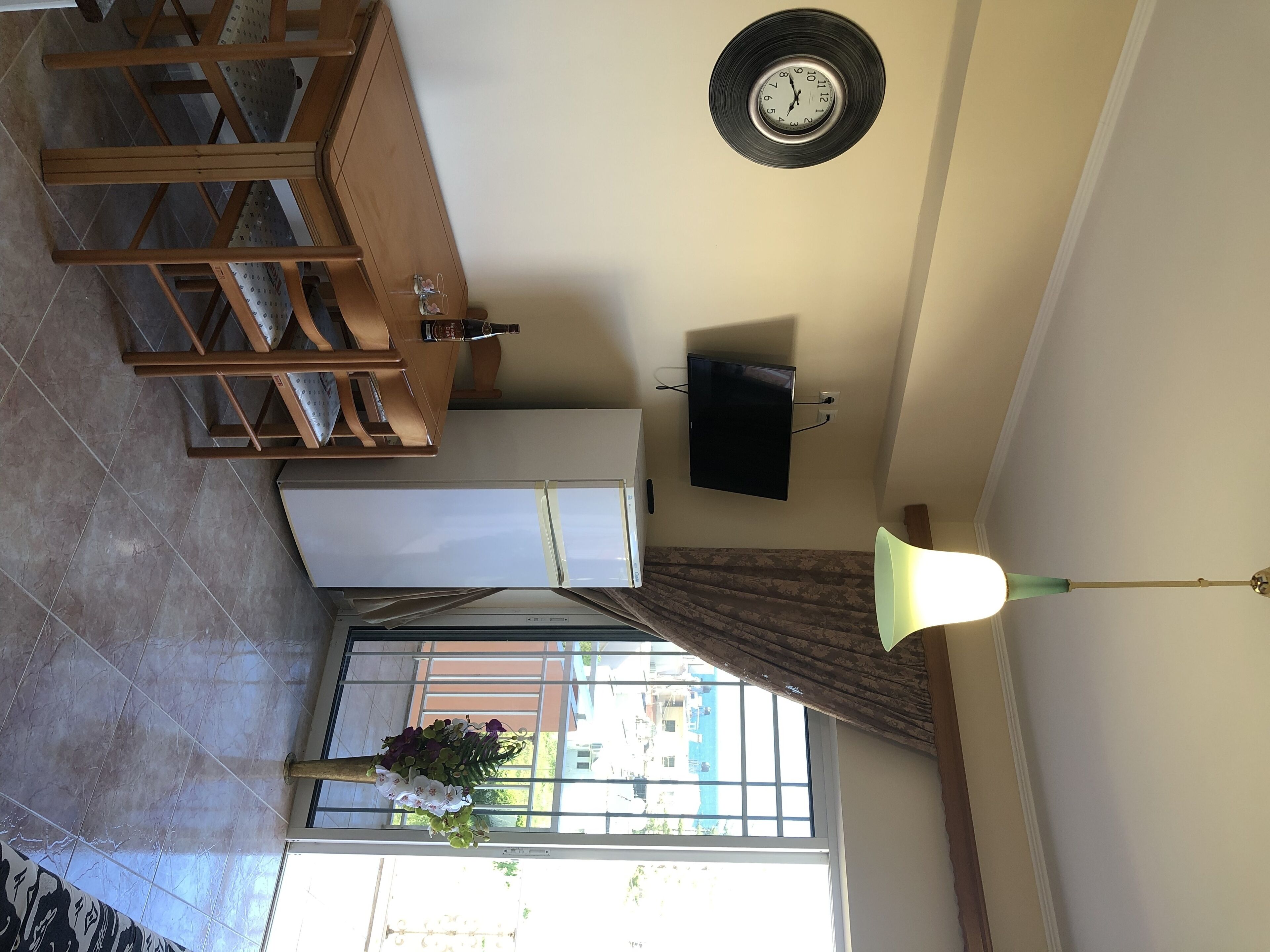 A room with a wooden dining table, a wall-mounted clock, a flat-screen TV, a ceiling fan, and a window with curtains.