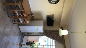 A room with a wooden dining table, a wall-mounted clock, a flat-screen TV, a ceiling fan, and a window with curtains. - Welcome to SEASIDE APARTMENT (agia marina)