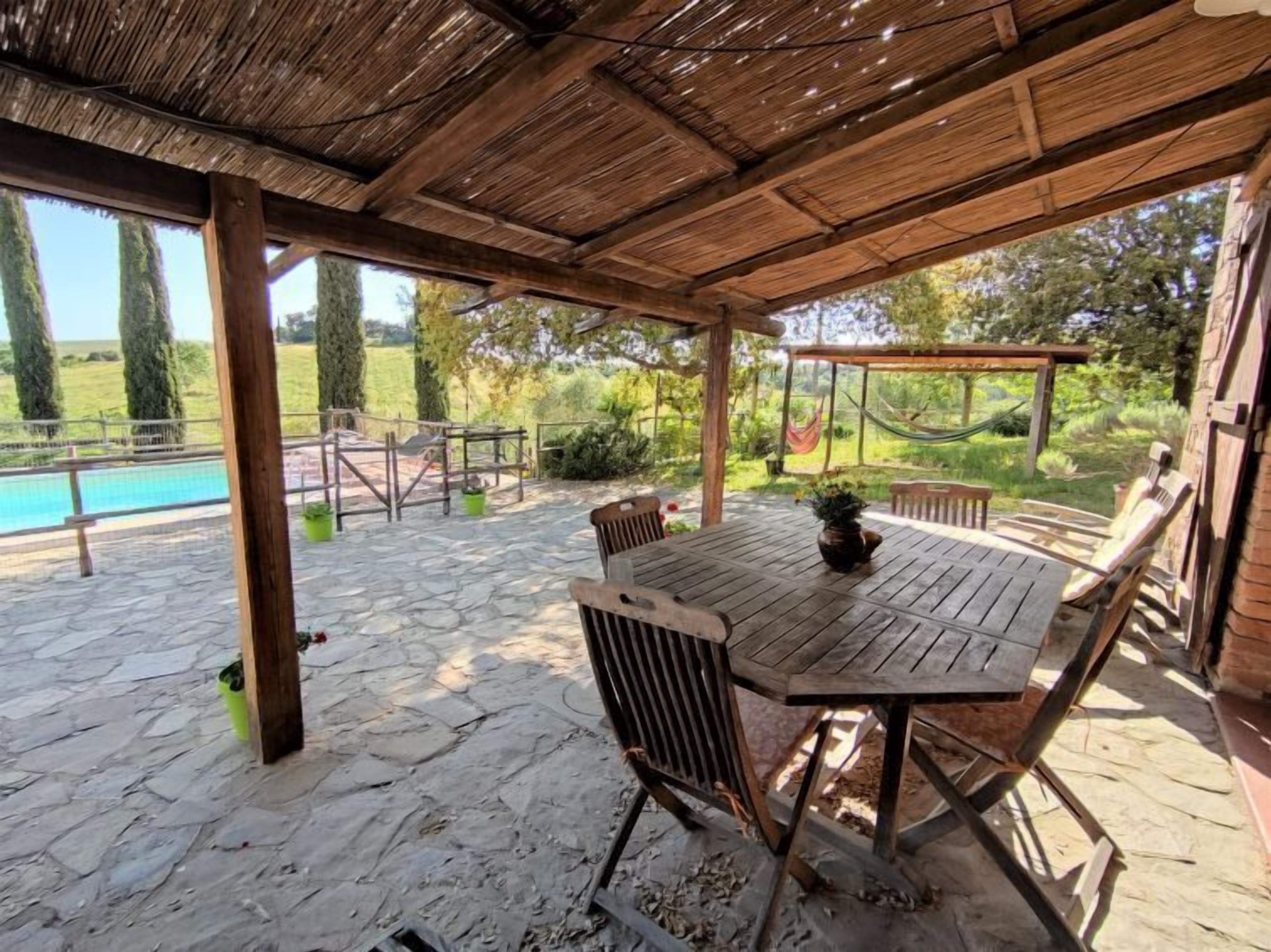 Un patio al aire libre con suelo de piedra, mesa y sillas de madera, una hamaca y vistas a una piscina y follaje.