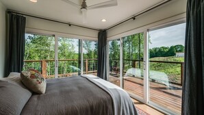 A bedroom with a large bed, a ceiling fan, and a view of a wooden deck and trees.