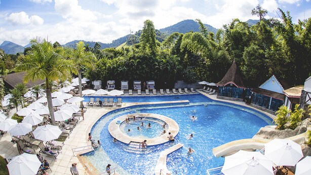 A large outdoor swimming pool with multiple lanes, surrounded by lounge chairs and umbrellas, set against a backdrop of lush greenery and mountains. - Magique Le Canton (Teresópolis)