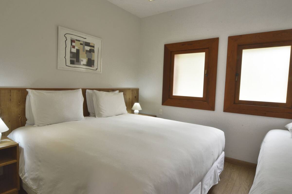 A hotel room with a large bed, two bedside tables with lamps, a wooden headboard, and two windows with wooden frames.