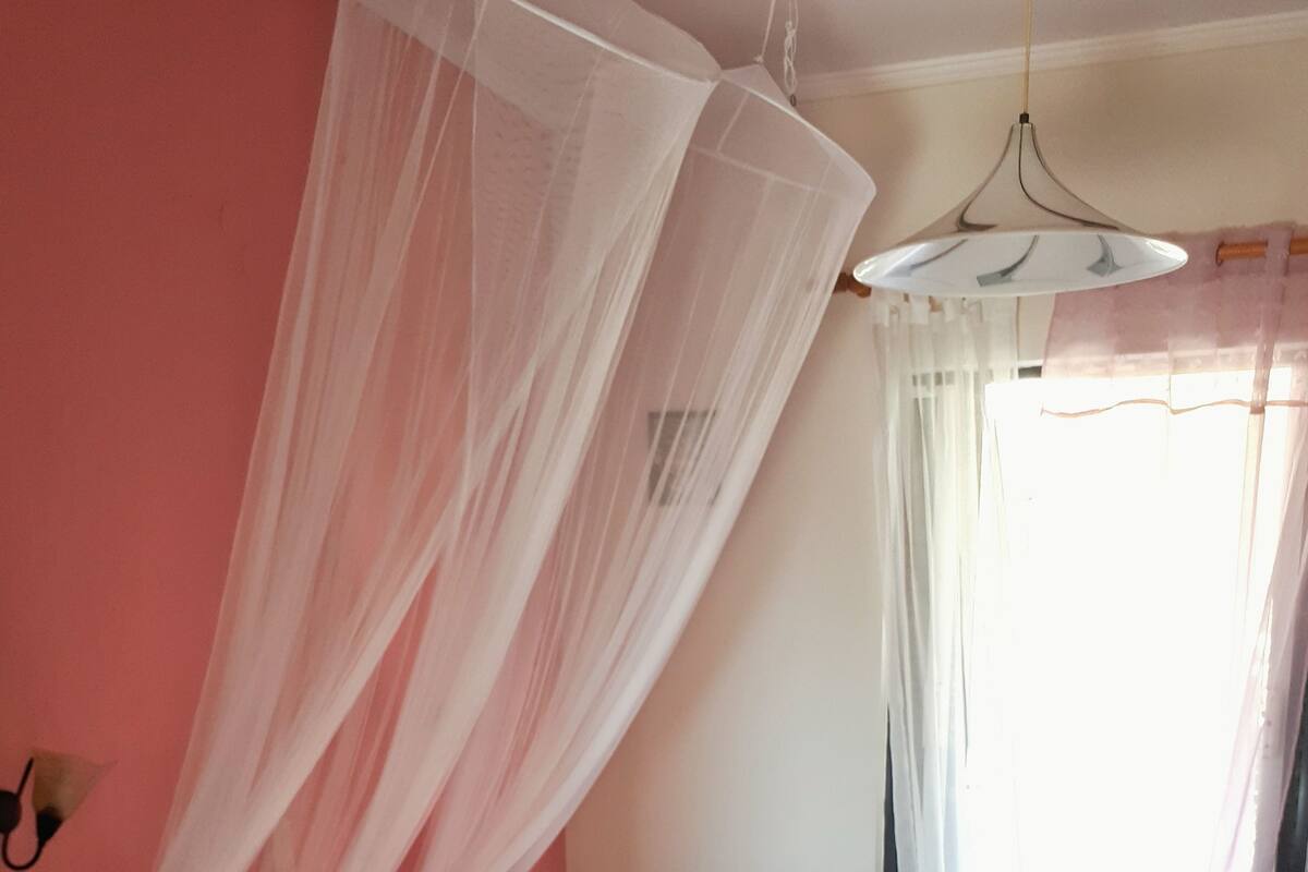 A room with two beds, a hanging light fixture, and sheer curtains.