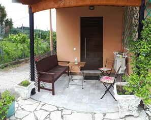 A covered outdoor seating area with a wooden bench, a small table, and a folding chair. - Antzela Apartments (Corfu)