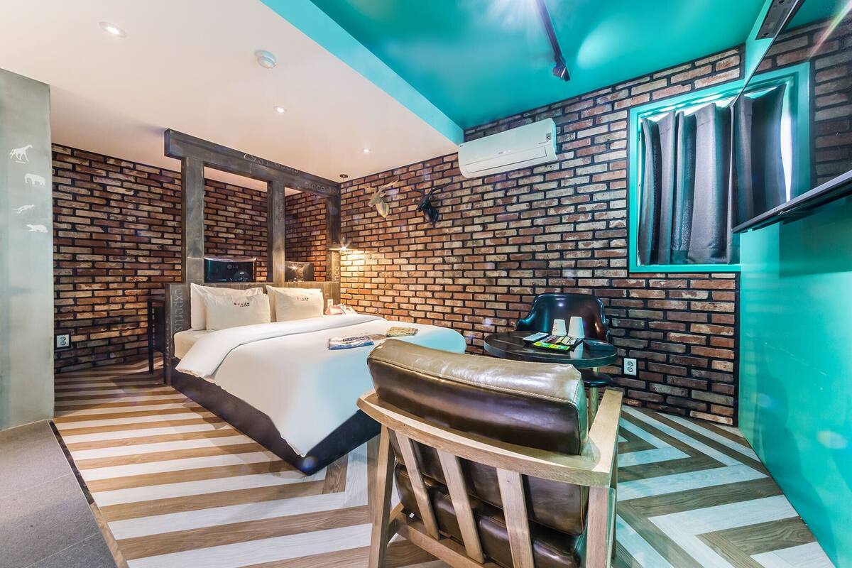 A modern hotel room with a brick wall, a bed with white linens, a leather armchair, a small table with a coffee machine, and a mounted TV.