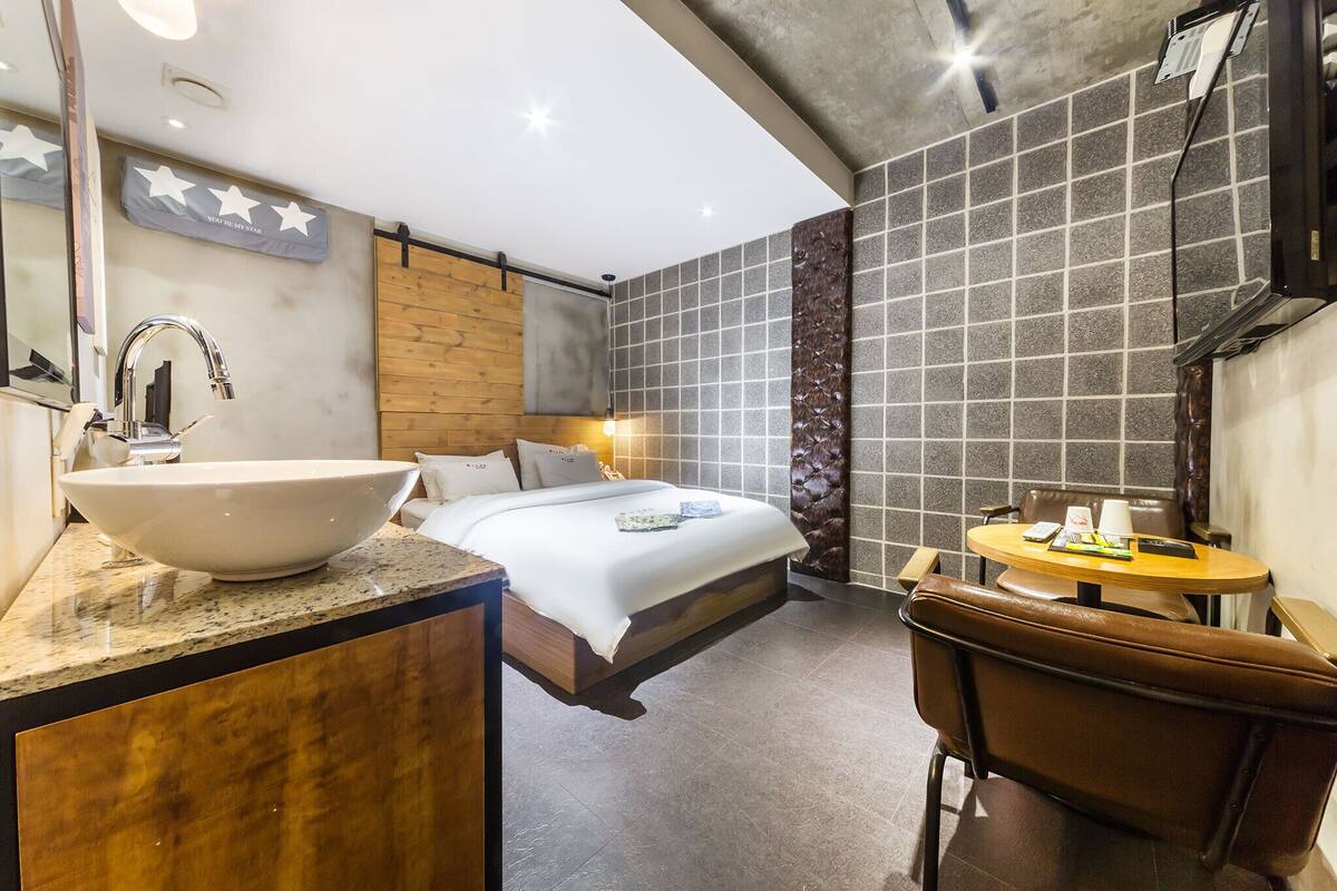 A hotel room with a large white sink, a bed with a wooden headboard, a round table with a chair, and a mirror on the wall.