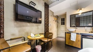 A modern kitchen with a large TV, a dining area with two chairs, a small round table, and a trash bin.