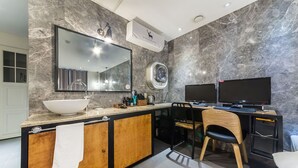 A modern bathroom with a large sink, mirror, and built-in desk with two monitors.