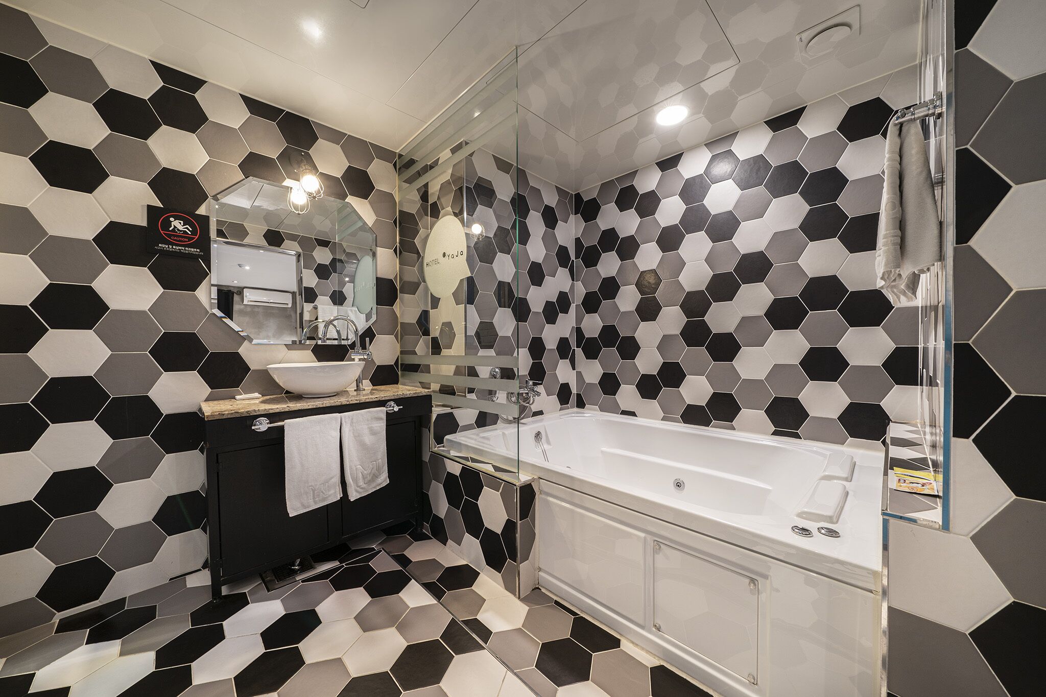 A modern bathroom with a black and white hexagonal tile pattern, a glass-enclosed shower, a white bathtub, and a black vanity with a mirror.