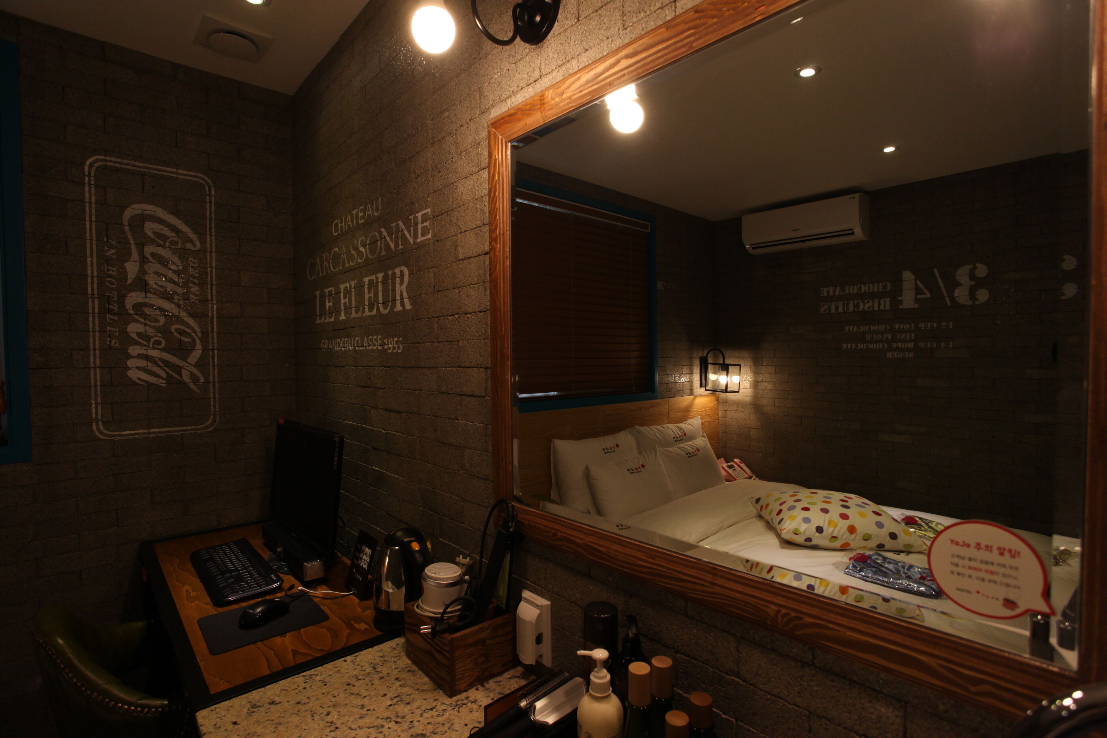 Une chambre d’hôtel avec un mur en briques, un bureau en bois avec un ordinateur, un lit avec des oreillers blancs à pois et un miroir qui reflète la pièce.