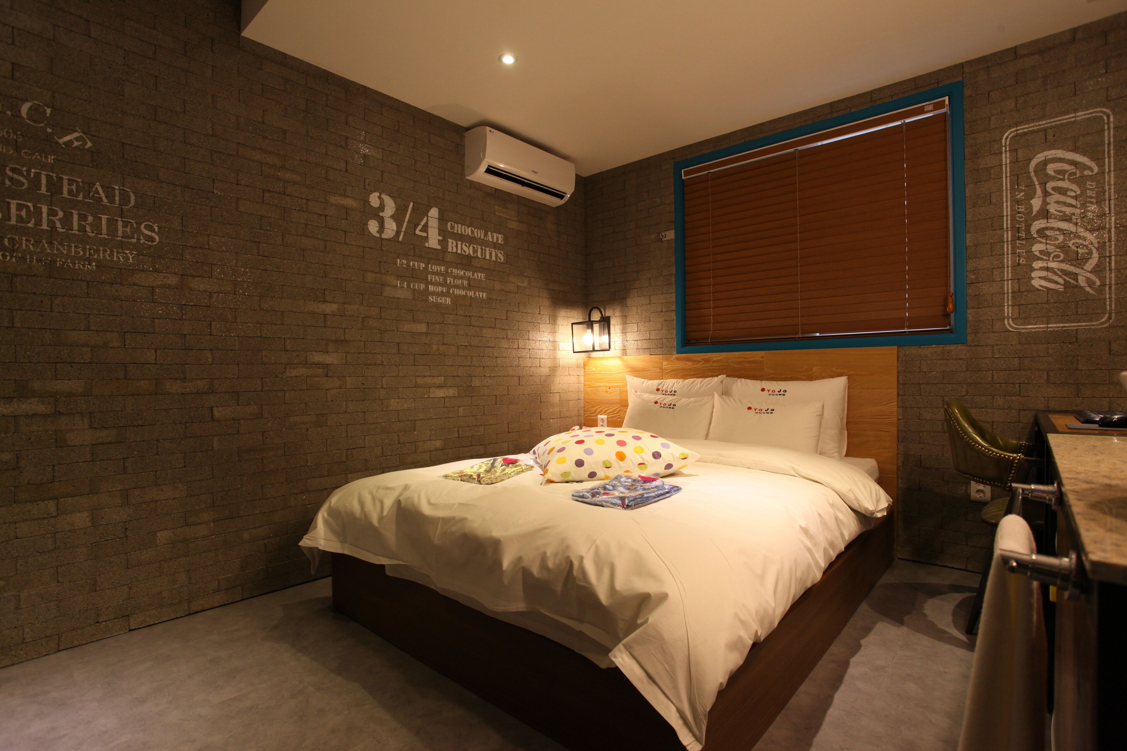 A hotel room with a brick wall, a bed with white bedding, a wooden headboard, a window with blinds, and a wall-mounted air conditioner.