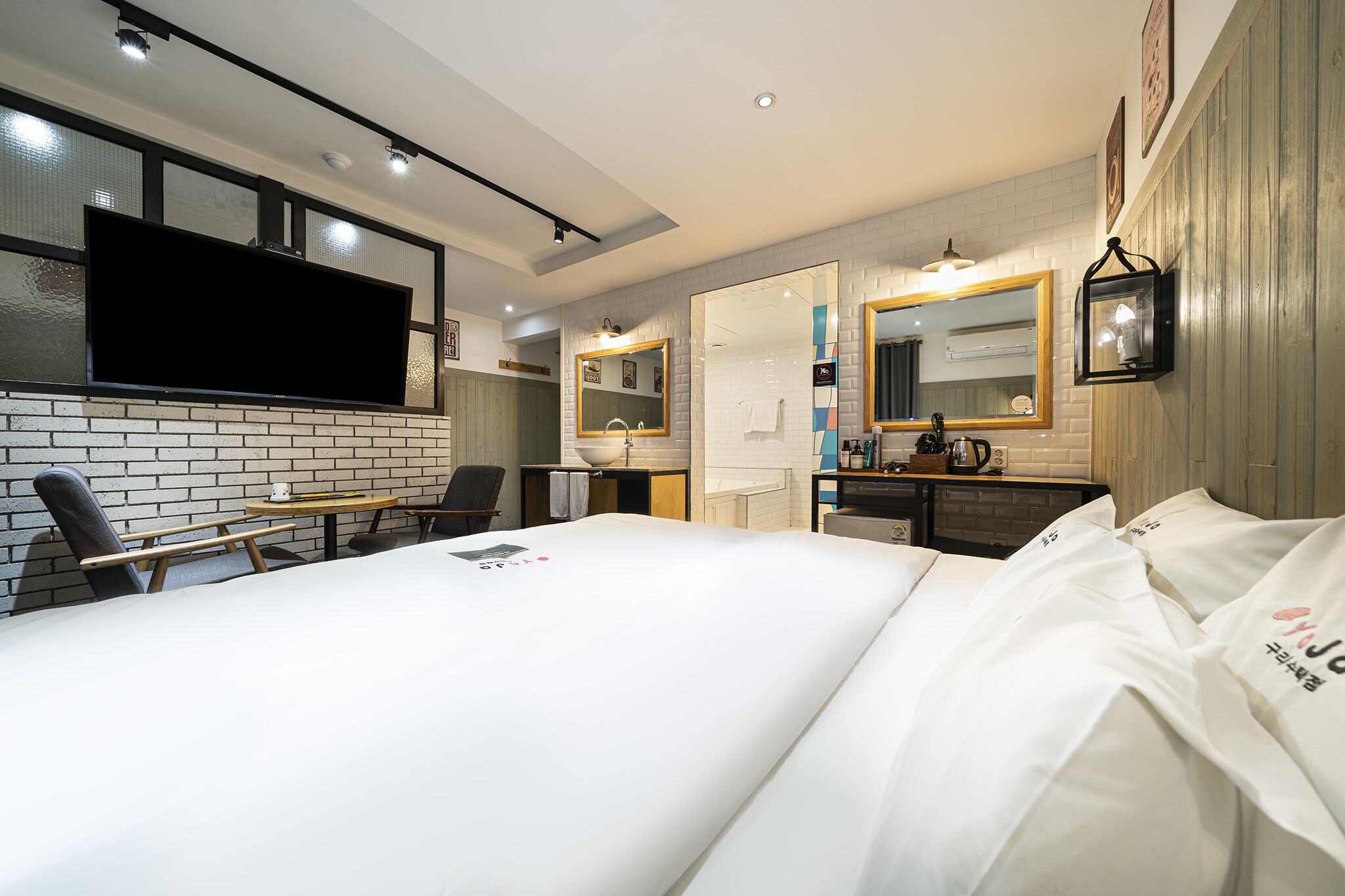 A modern hotel room with a large bed, a flat-screen TV mounted on the wall, a small desk with a chair, and a bathroom with a mirror and sink.