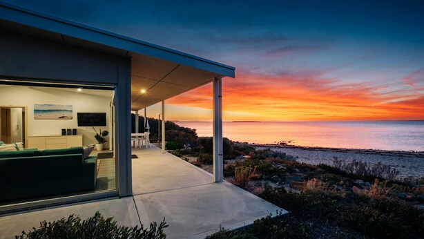 A modern house with a covered outdoor area, a view of the sea at sunset, and a dining area with a table and chairs.