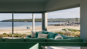 A room with a large window overlooking a beach, a green sofa, and a view of two people walking on the shore.