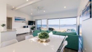 A modern living room with a large window overlooking a beach, a dining area with a white table, and a green sofa.