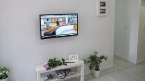 A television mounted on a white wall displaying a news program. Below the TV, there is a white shelf with a potted plant, a framed picture, and some magazines.