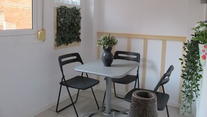 A small dining area with a table and four chairs, a hanging light fixture, a wall-mounted plant, and a window with a view of a brick building.