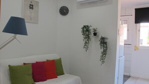 A white sofa with colorful cushions, a wall clock, an air conditioning unit, and hanging plants.