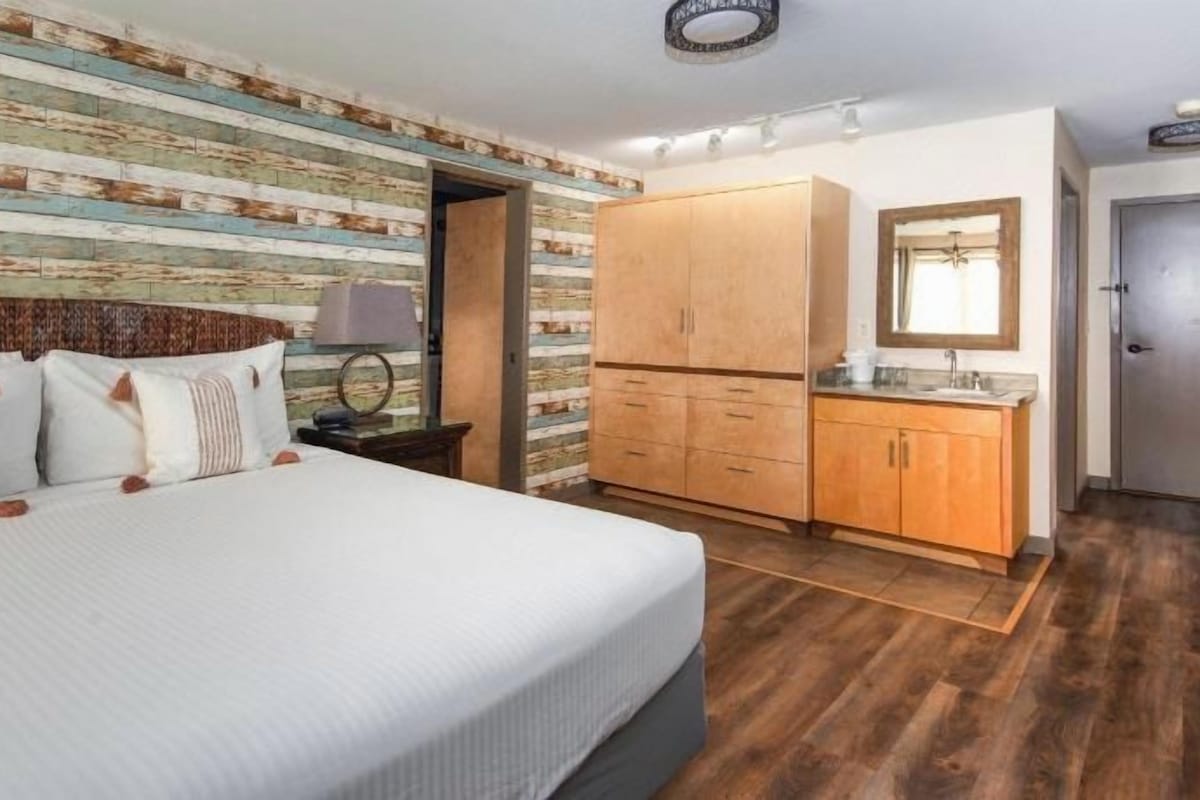 A bedroom with a large bed, wooden wardrobe, and a small bathroom with a mirror and sink.
