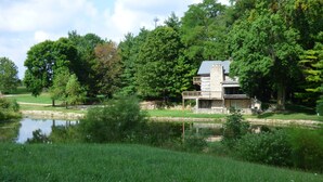 A stone building with a balcony, surrounded by lush greenery and a pond. - Beautiful, 200 year old Log home - Secluded, 30 Min. from Downtown (Indianapolis, Acton)
