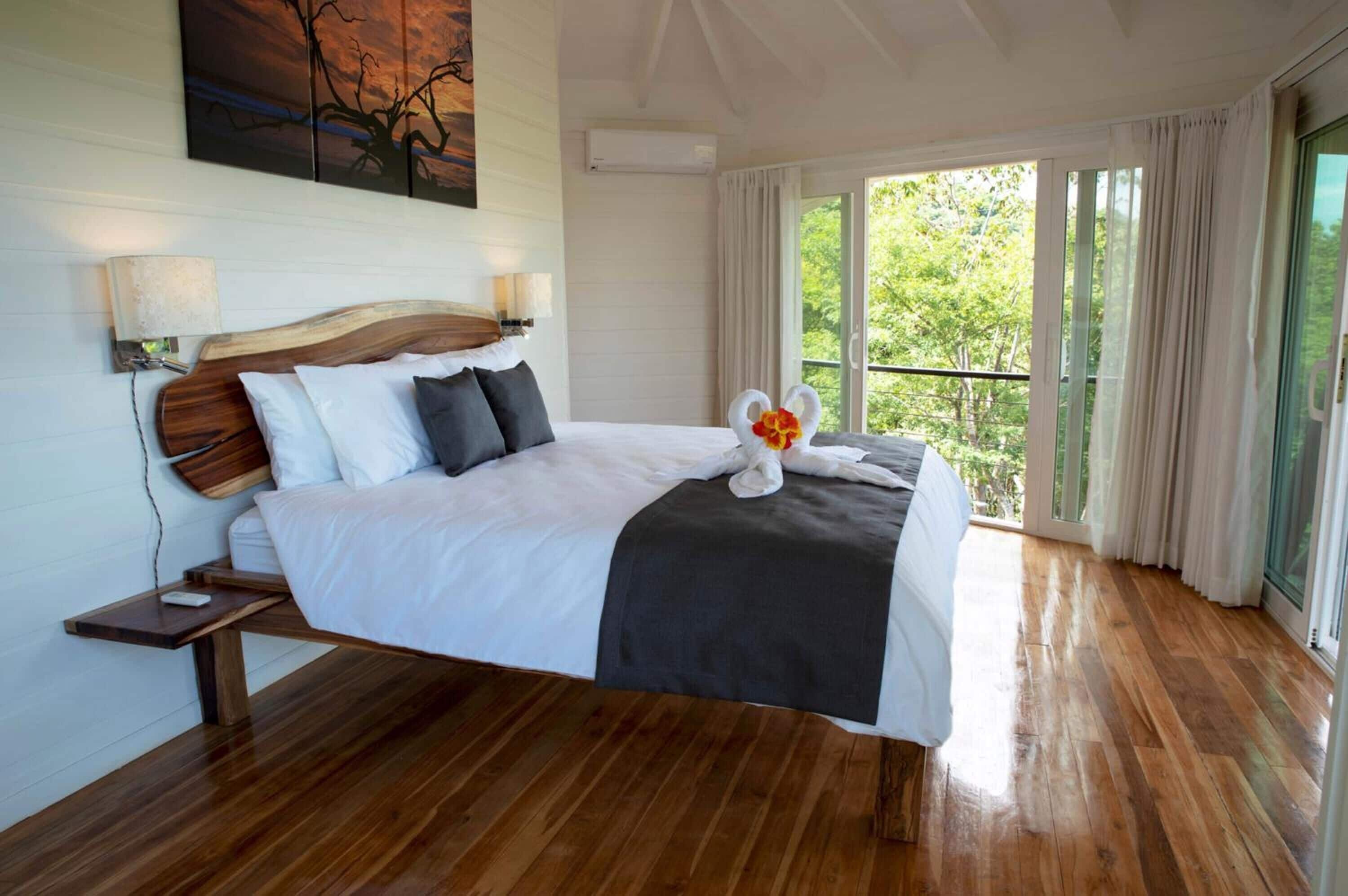 A large bed with a wooden headboard, white bedding, and a decorative swan pillow. There is a nightstand with a lamp and a small table beside the bed. The room has wooden flooring and large windows with curtains.