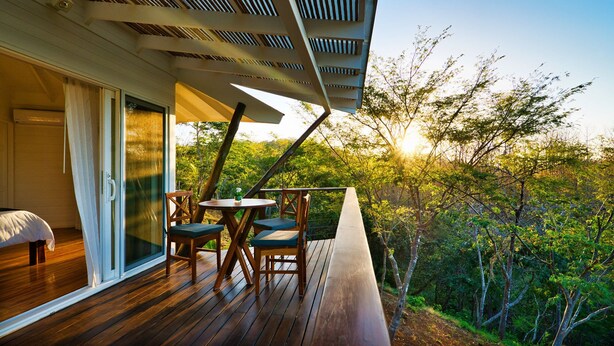 A wooden deck with a table and chairs, a sliding glass door leading to a bedroom, and a view of a forested area. - Mikado Natural Lodge (Tamarindo)