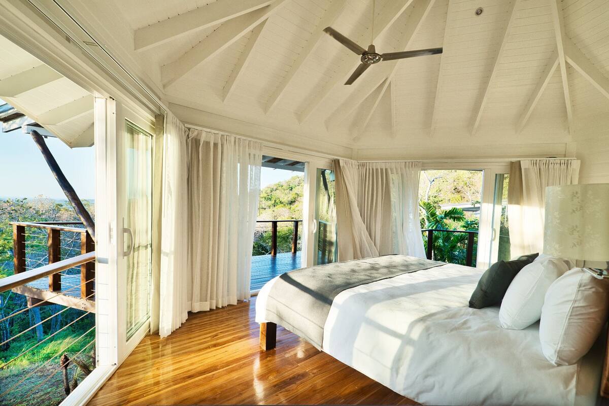 A bedroom with a bed, a ceiling fan, and large windows offering a view of the outdoors.