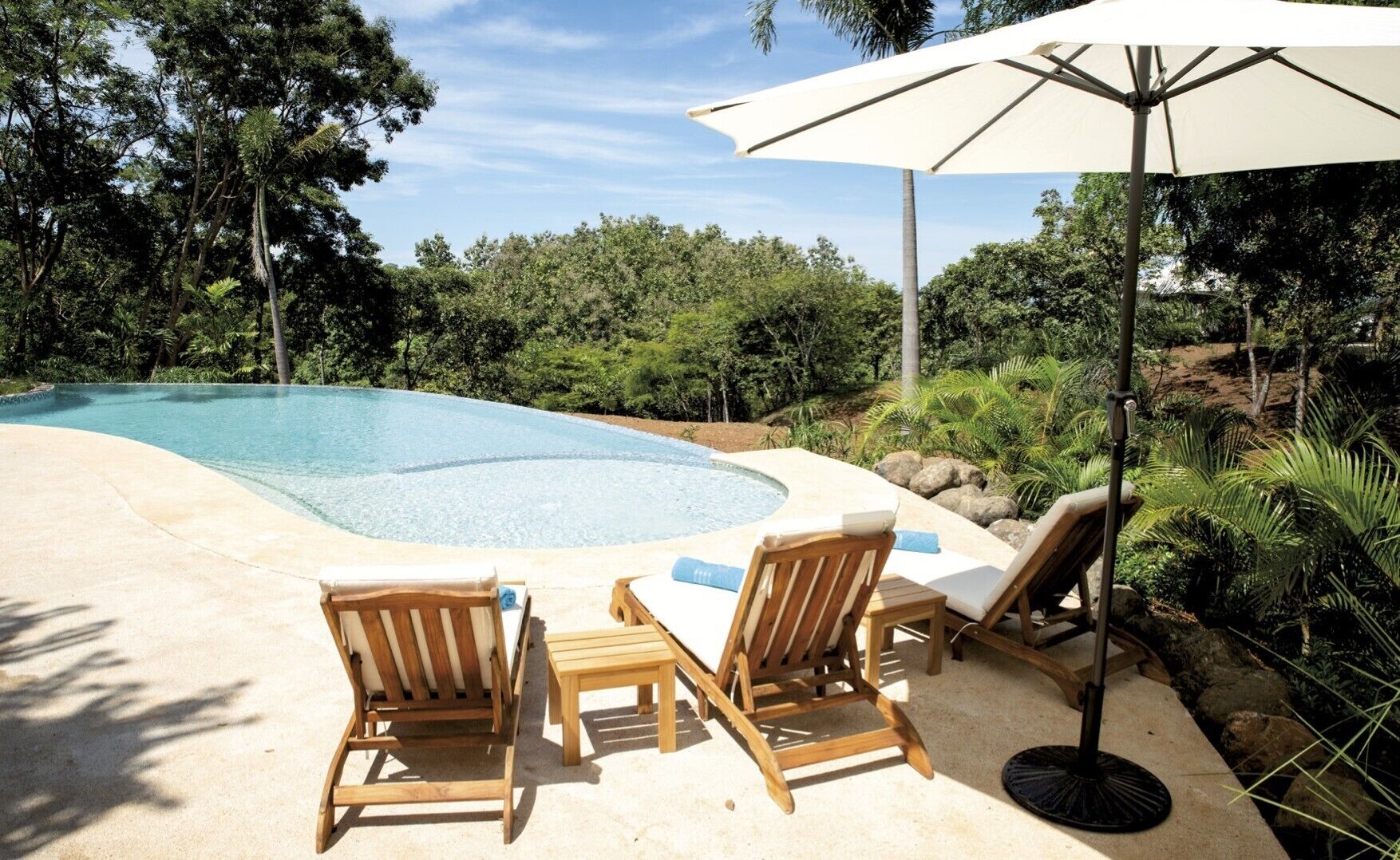 A poolside area with lounge chairs and a table, surrounded by lush greenery and a clear blue pool.