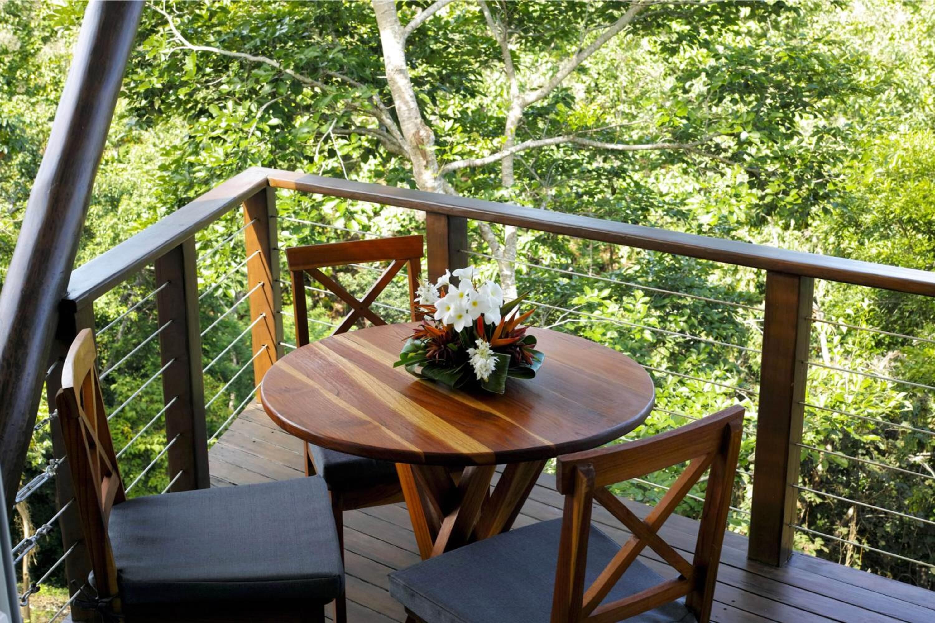 A wooden deck with a round table and two chairs, surrounded by a metal railing, overlooking a lush green landscape.