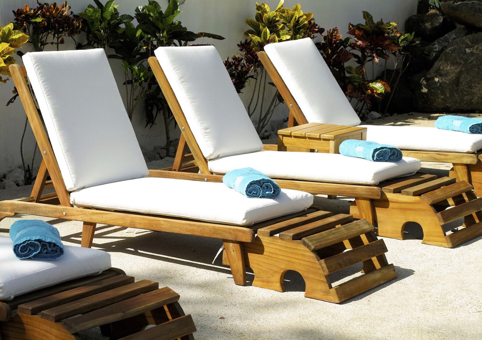A row of wooden lounge chairs with white cushions and blue towels, arranged on a sandy surface with plants in the background.
