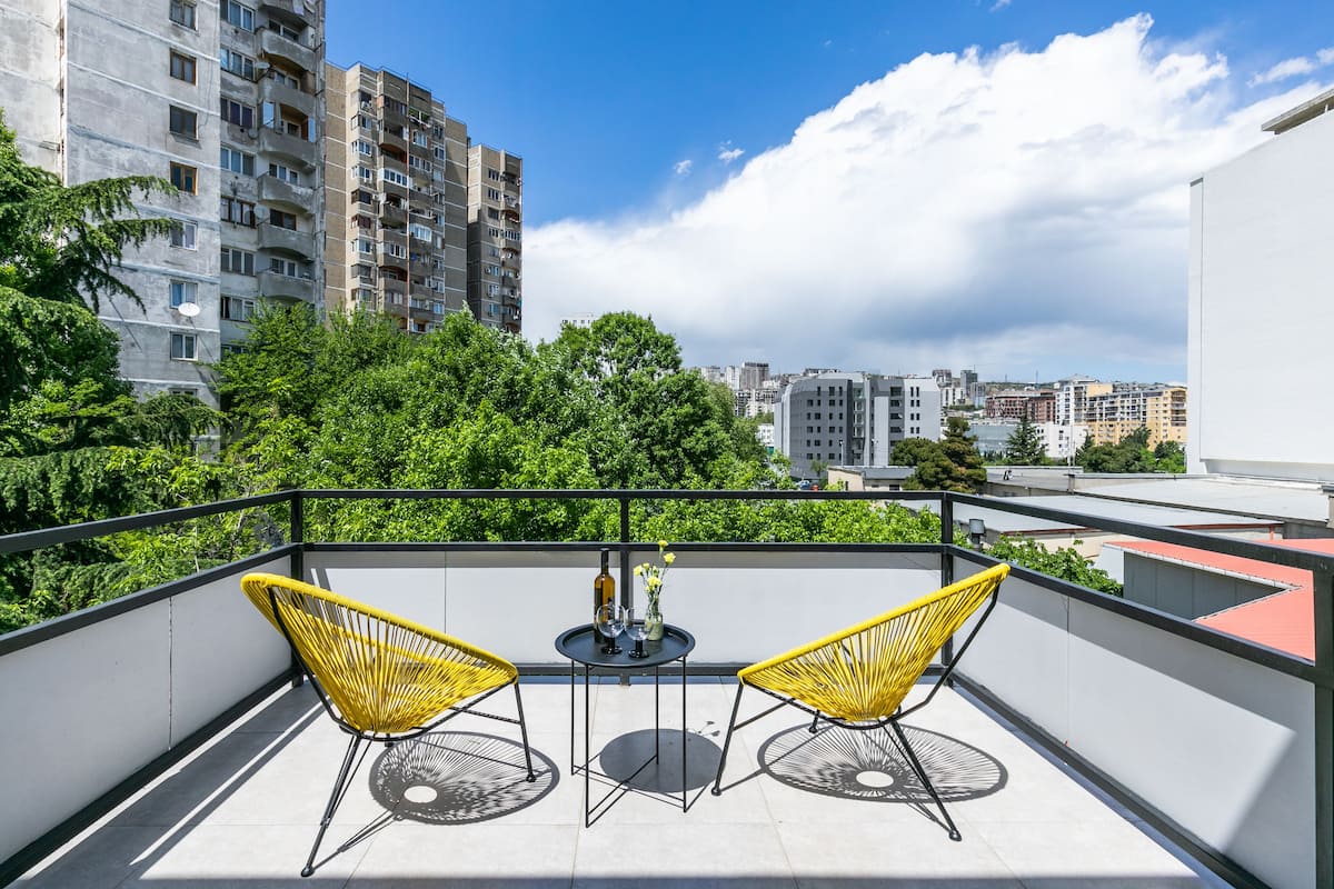 A balcony with two yellow chairs, a small table with a bottle and flowers, and a cityscape view.