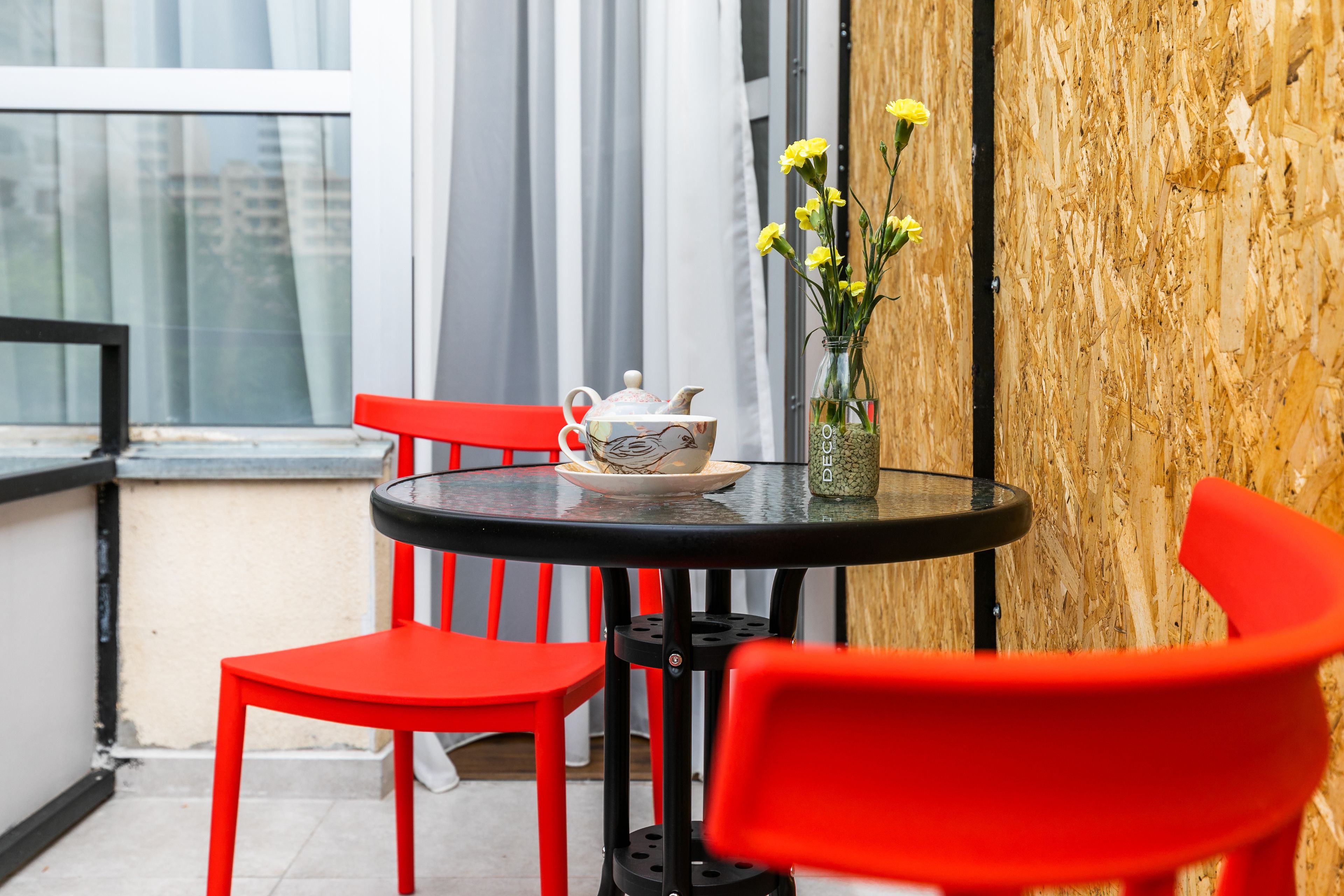 A small round table with a teapot and a vase of yellow flowers, red chairs, and a wooden wall.