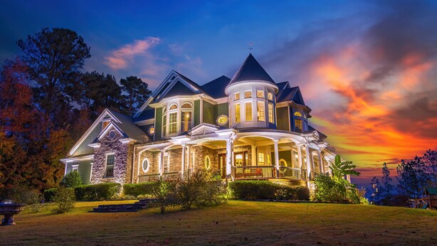 Une grande maison à étages, dotée d’une véranda qui fait le tour de la maison et d’une tourelle, et entourée de pelouses et d’arbres bien entretenus.