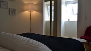 Quarto com cama, luminária, cadeira vermelha e janela com vista para um edifício.