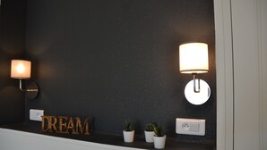 Parede com superfície escura e texturizada, duas luminárias fixadas na parede, uma tomada e três pequenos vasos com plantas.