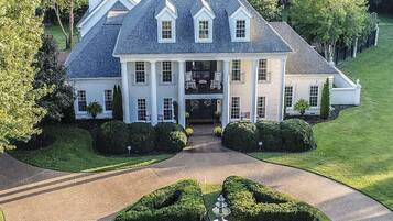 A large, white, two-story house with a symmetrical facade, a central entrance, and a formal garden with trimmed hedges and a fountain.