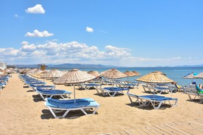 A beach with rows of sun loungers and thatched umbrellas, a wooden boardwalk, and a clear view of the sea and distant mountains. - Otel Türk Evi (Ayvalik)
