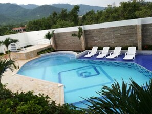 A swimming pool with white lounge chairs, surrounded by a stone wall and greenery, with mountains in the background.
