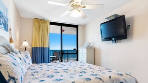 Habitación de hotel con cama, ventilador de techo, televisor montado en la pared y vista al océano por la ventana.