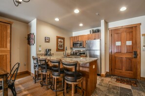A kitchen with wooden cabinets, a stainless steel refrigerator, a microwave, and a bar area with stools.