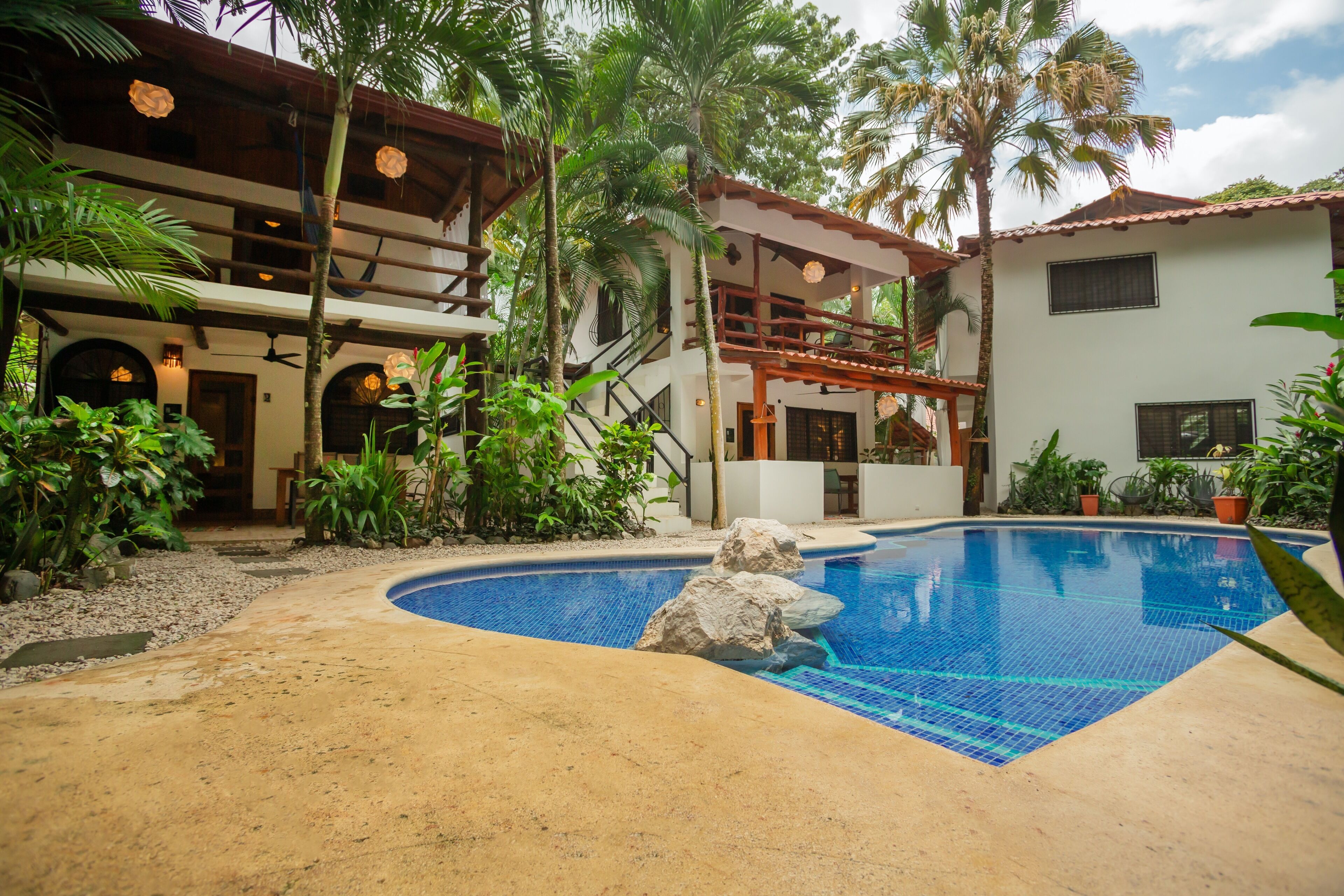 A pool area with a clear blue pool, surrounded by palm trees and a two-story building with balconies.