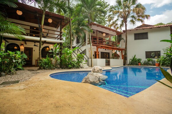 A pool area with a clear blue pool, surrounded by palm trees and a two-story building with balconies.