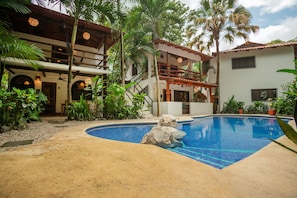 A pool area with a clear blue pool, surrounded by palm trees and a two-story building with balconies. - Villa Palma, Live outside and sleep in the AC at Villa Palma (Sámara)