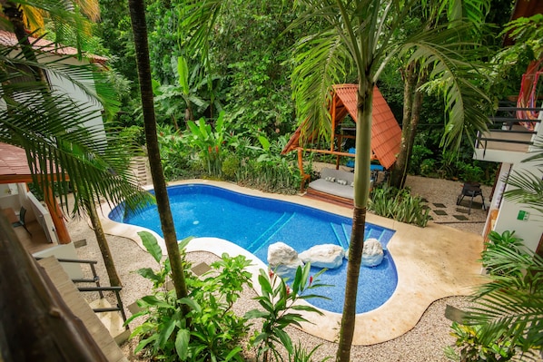 A swimming pool surrounded by tropical vegetation, with a wooden deck and a small structure with a red roof.