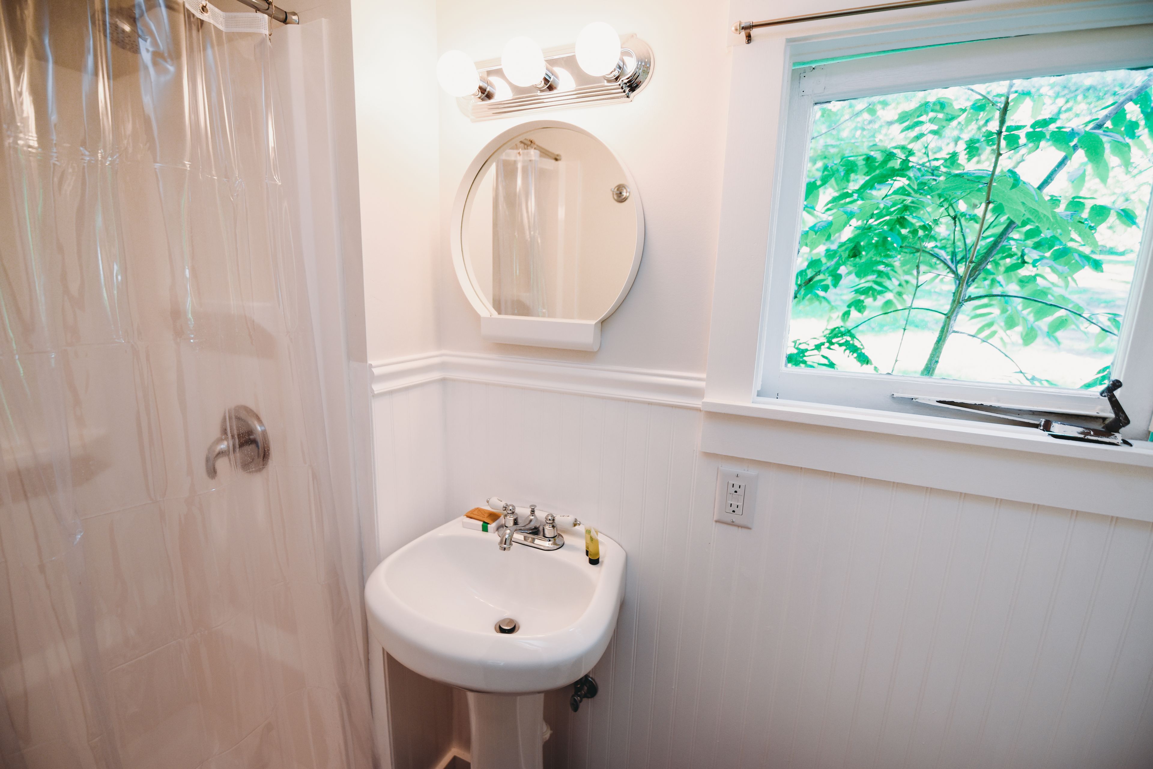 Un bagno con tenda da doccia, lavandino con specchio sopra e finestra vista alberi.
