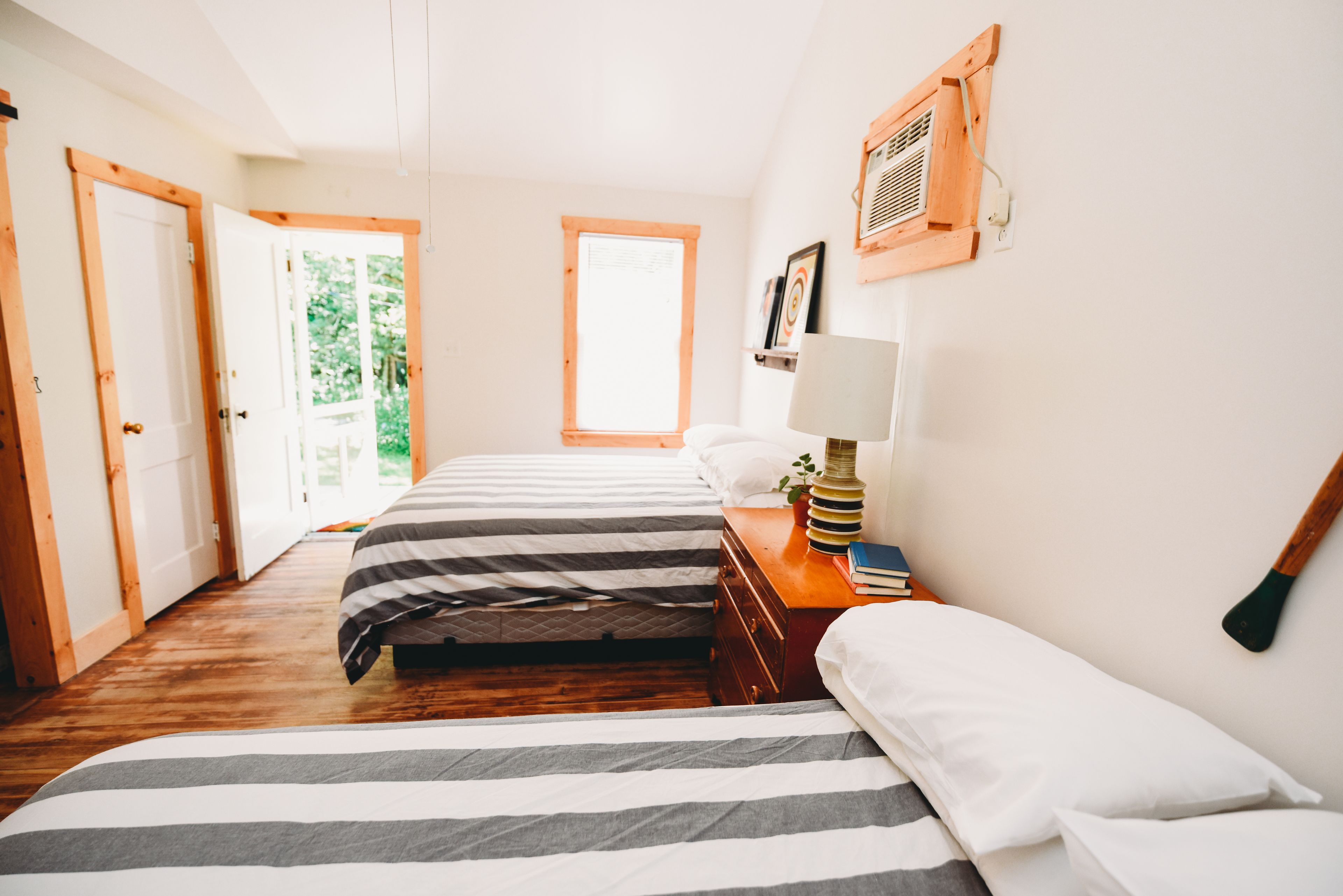 Un dormitorio con dos camas, piso de madera, una mesita de noche con libros, una lámpara y una ventana con vista a la vegetación.