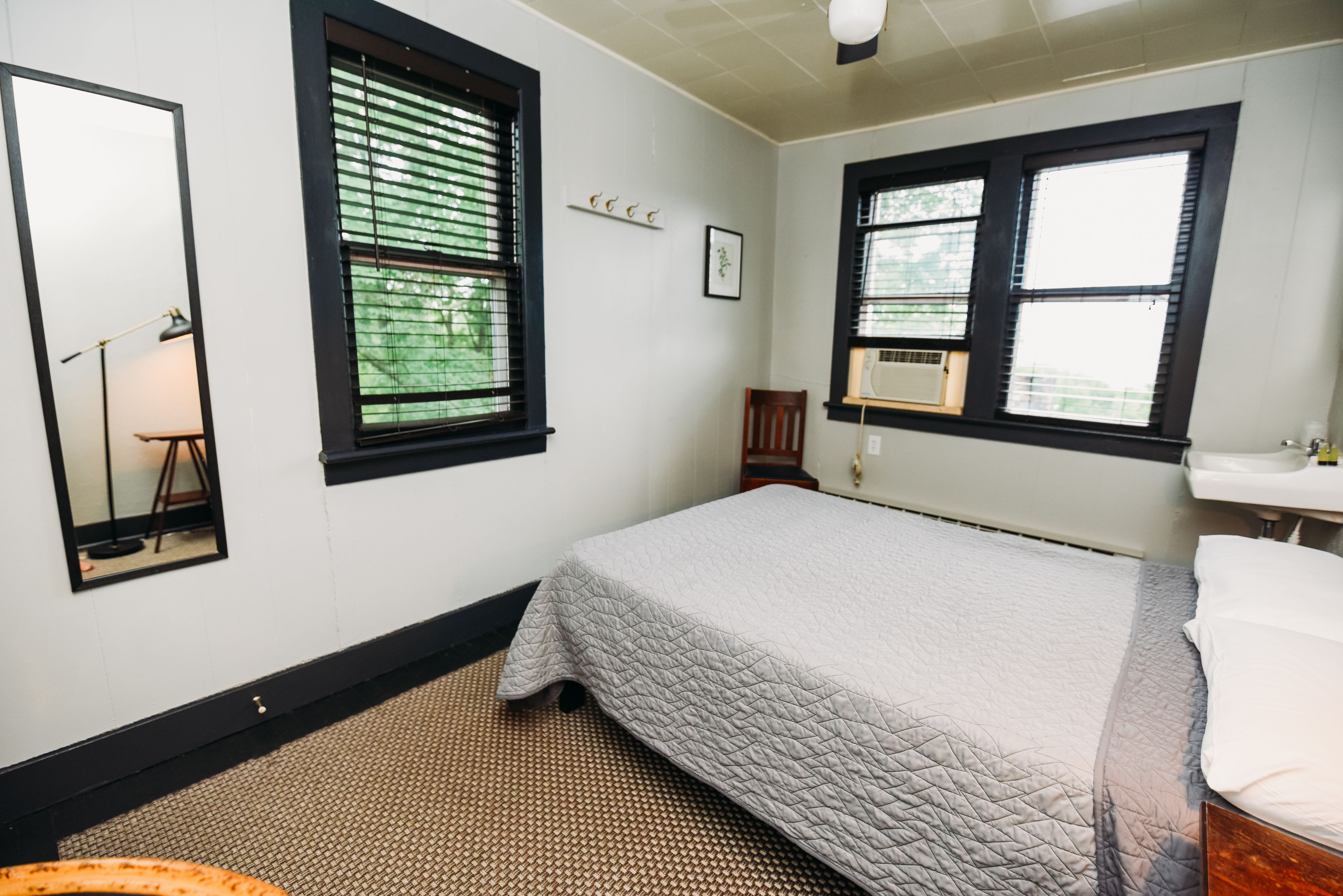 A bedroom with a bed, a mirror, a window with blinds, a radiator, and a small wooden chair.
