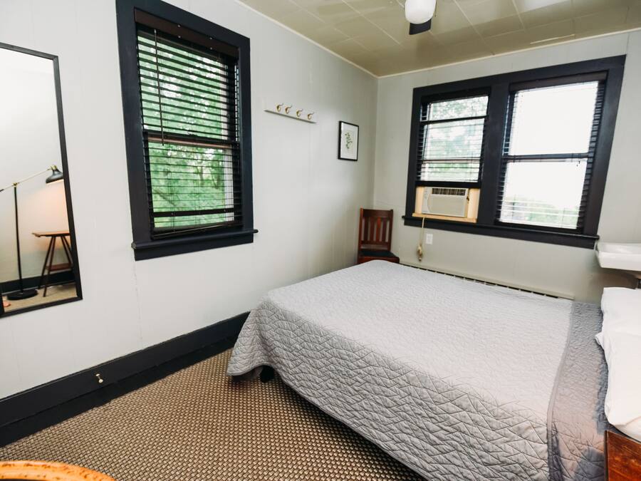 Un dormitorio con cama, espejo, ventana con persianas, radiador y una pequeña silla de madera.